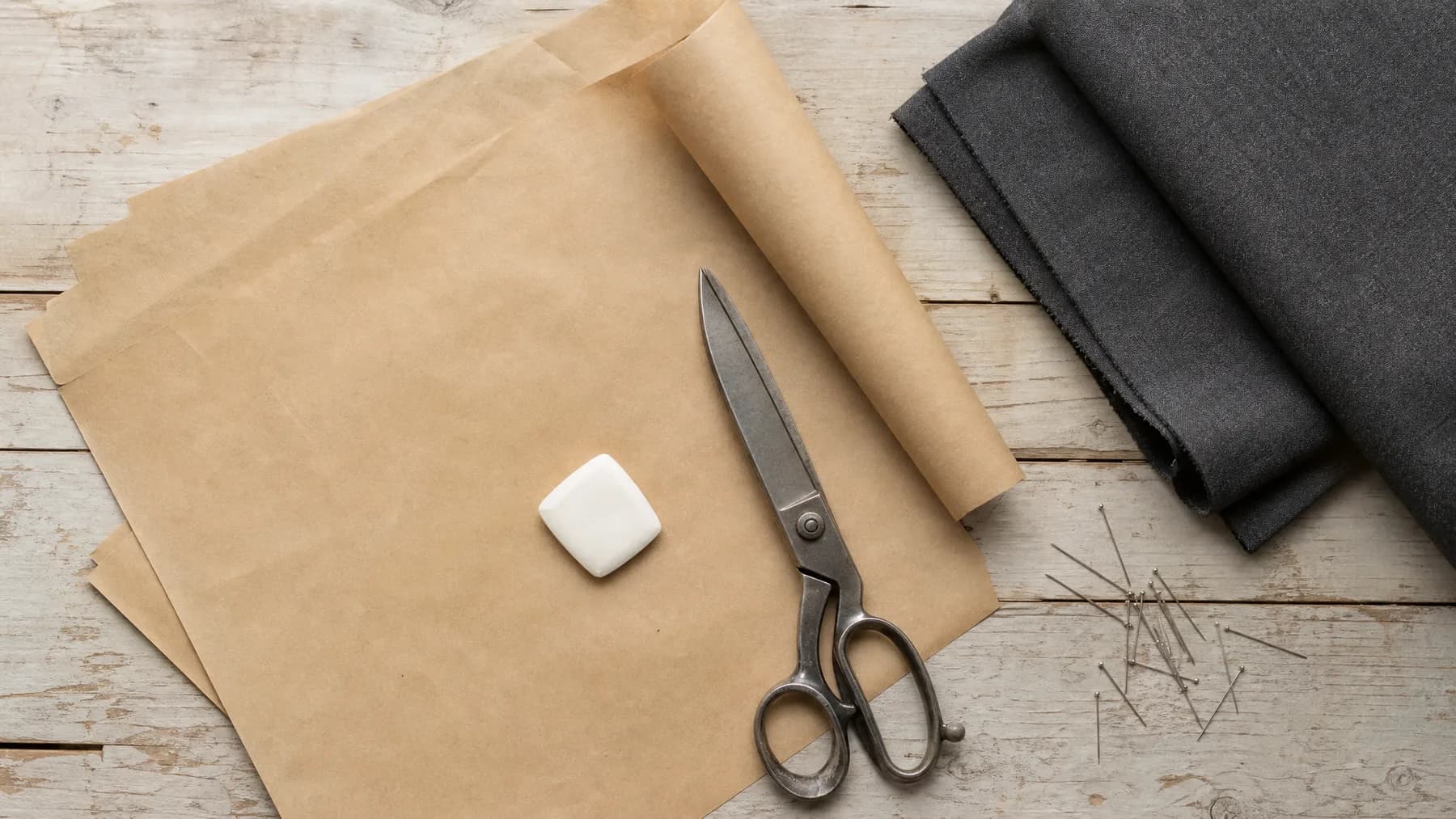 A tailor's cutting table seen from above: kraft pattern paper, chalk, shears, folded charcoal cloth and pins.