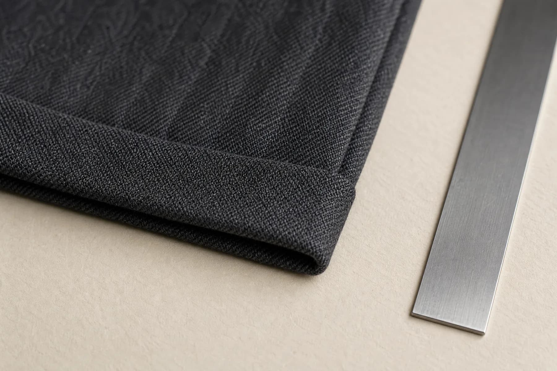 Extreme close detail of the pressed bottom hem of a charcoal wool trouser leg lying on pale paper beside a thin steel rule.