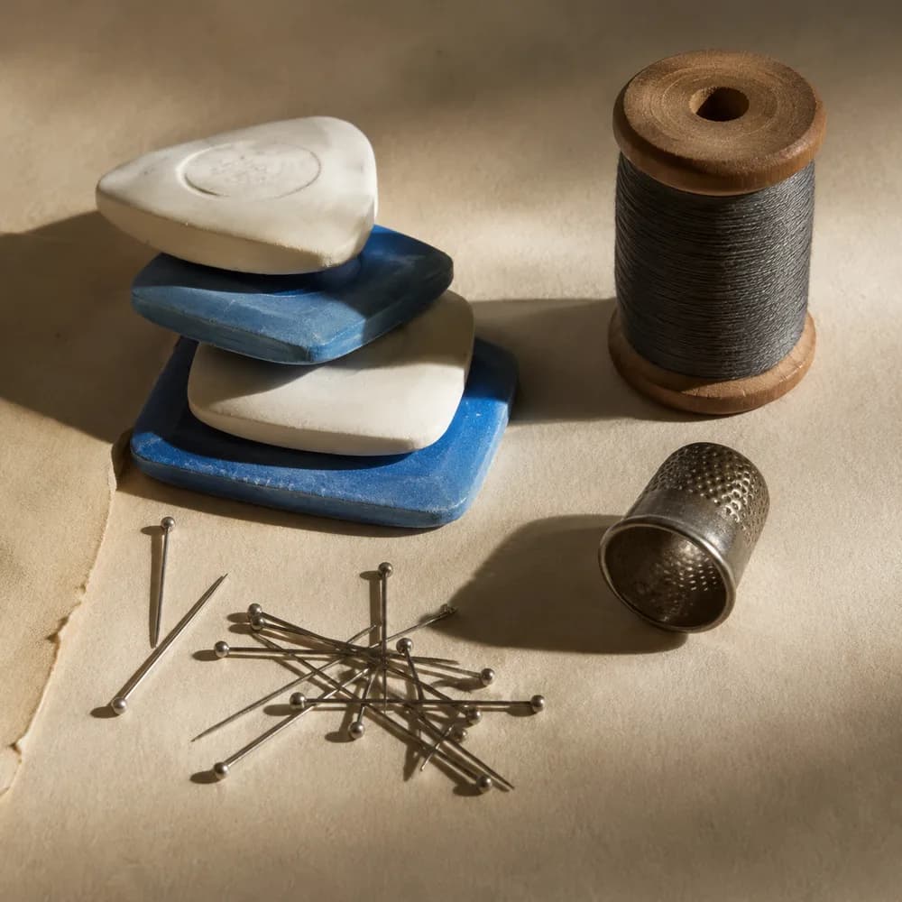 Tailor's chalk, steel pins, a spool of dark thread and a thimble on cream paper.